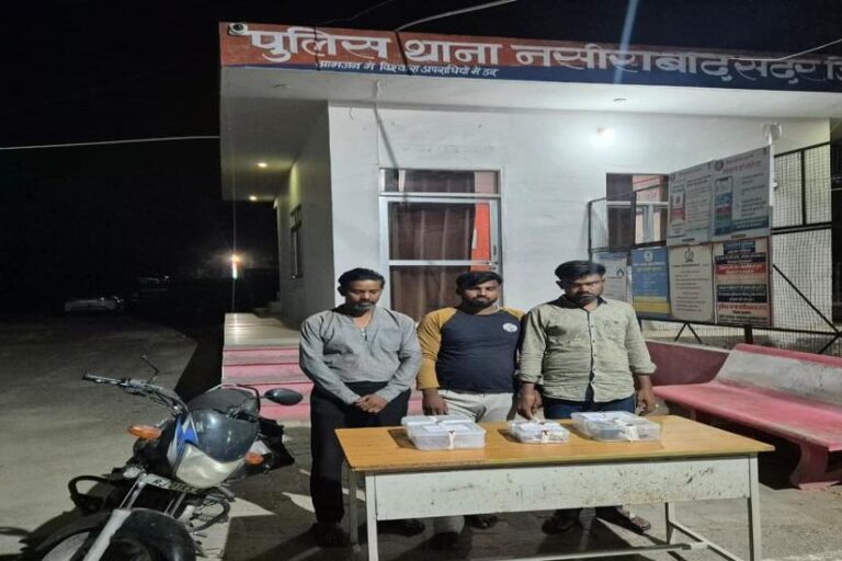 Three accused standing behind a table of recovered jewellery in front of Nasirabad Sadar police station, with a motorcycle beside them